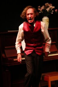 The actor as Chopin standing before an upright piano mid-speech, flowers in a vase behind him