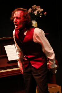 Chopin leaning forward beside the piano, sheet music on the stand, speaking intently