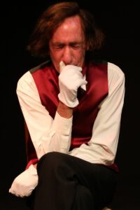 Chopin seated, raising a white-gloved hand to his mouth as he coughs