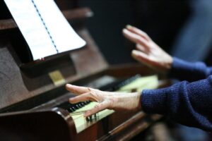 White-gloved hands at the keyboard of an upright piano, sheet music above