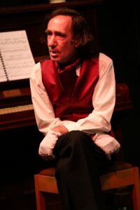 Chopin seated on a chair beside the piano, legs crossed and hands clasped