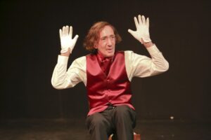 Chopin seated on a darkened stage with both gloved hands raised