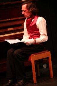 Chopin seated on a stool beside the piano, reading sheet music