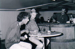 Two men in conversation at a pub table while the barman works behind the bar